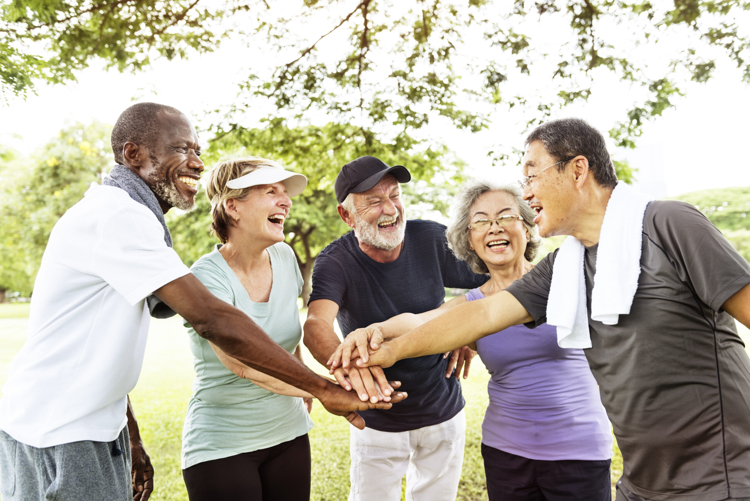 Diverse group of five smiling older adults in workout clothes standing in a half circle in a park with their hands stacked in the center of the group